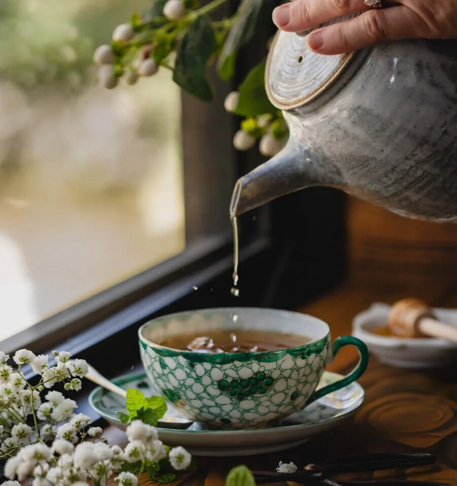 Tea being poured from a silver teapot into a green and white patterned cup on a wooden table with white flowers nearby.
