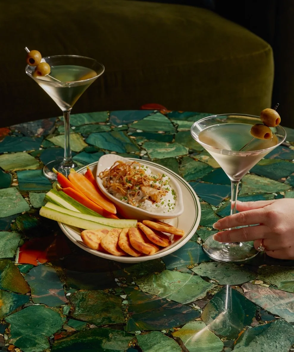 Two martini glasses with olives on a colorful, leaf-patterned table, alongside a plate of sliced vegetables, crackers, a dip, and a small bowl of salad.