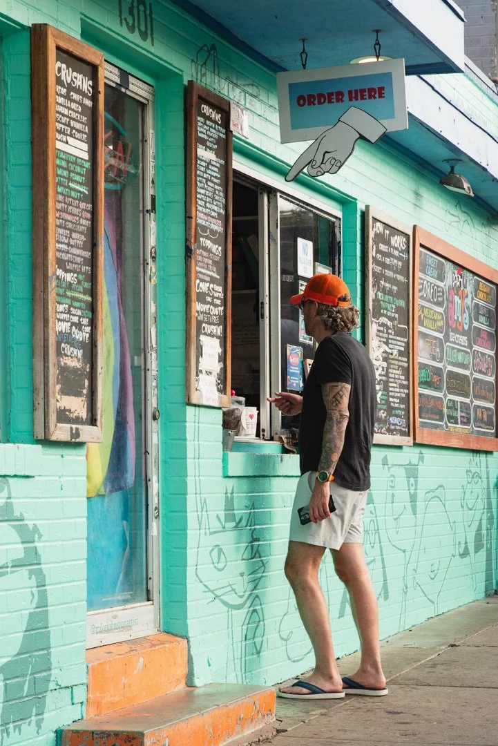 A person in sunglasses and a red cap orders ice cream at a colorful ice cream shop with multiple chalkboard menus and an 'Order Here' sign overhead.