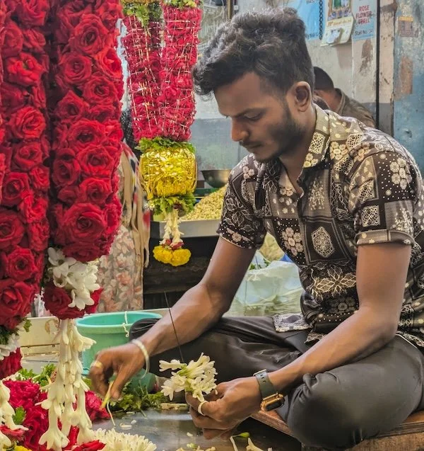 A man tying white flower garlands in a flower shop with vibrant red, yellow, and white flowers around him.