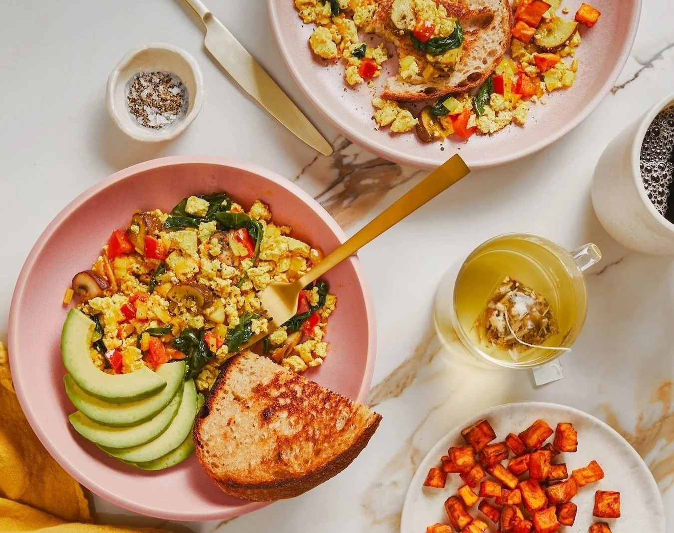A breakfast spread with two pink bowls of scrambled eggs mixed with vegetables, served with avocado slices, a slice of toasted bread, and a fork. There is a white plate of small diced roasted sweet potatoes, a cup of black coffee, a glass mug of herb