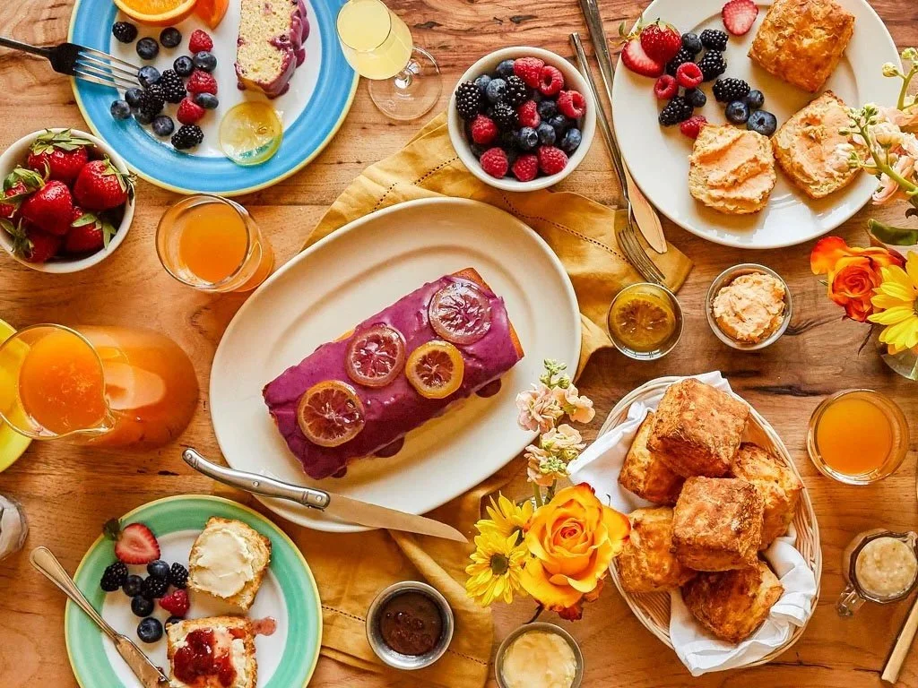 A colorful breakfast or brunch spread on a wooden table, featuring various plates of pastries, fruit, and beverages. There are bowls of strawberries, blueberries, and raspberries, a large purple roulade cake garnished with blood orange slices, and se