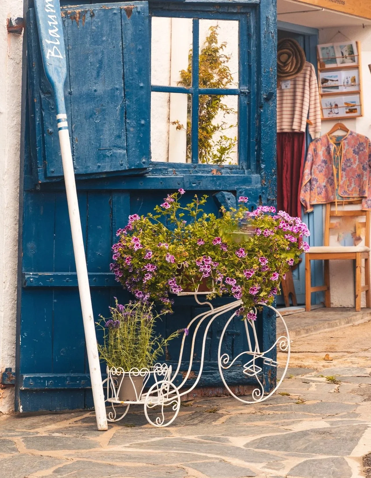 A rustic scene featuring a blue wooden window shutter with a small open window, a blooming pink and purple flower pot on a decorative white metal bicycle stand, and a cozy outdoor shop with hanging clothes and framed photos in the background.
