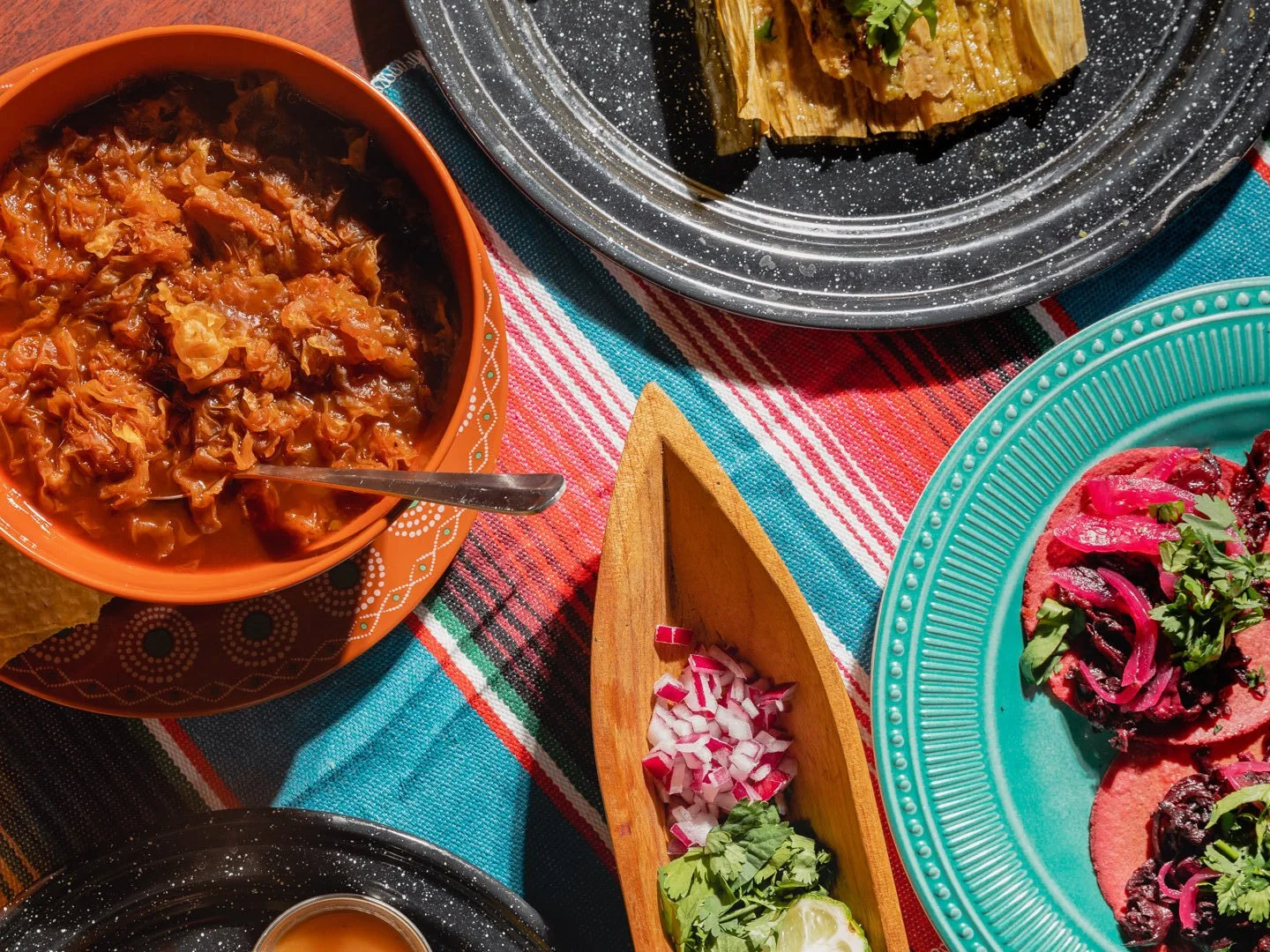 Close-up of Mexican food dishes on colorful tablecloth, including a bowl of shredded meat in sauce, a platter of tacos topped with radish, cilantro, and onions, and a plate of tamales garnished with greens.