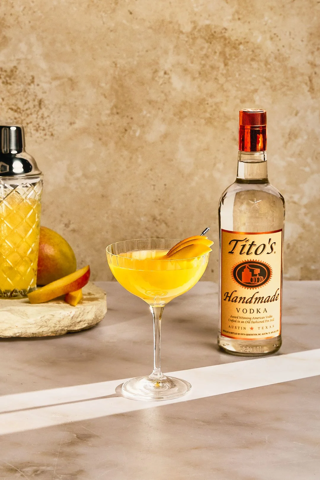 A cocktail glass with a yellow drink and lemon garnish, a bottle of Tito's Handmade Vodka, and a fruit tray with sliced apples and whole fruits on a marble surface.