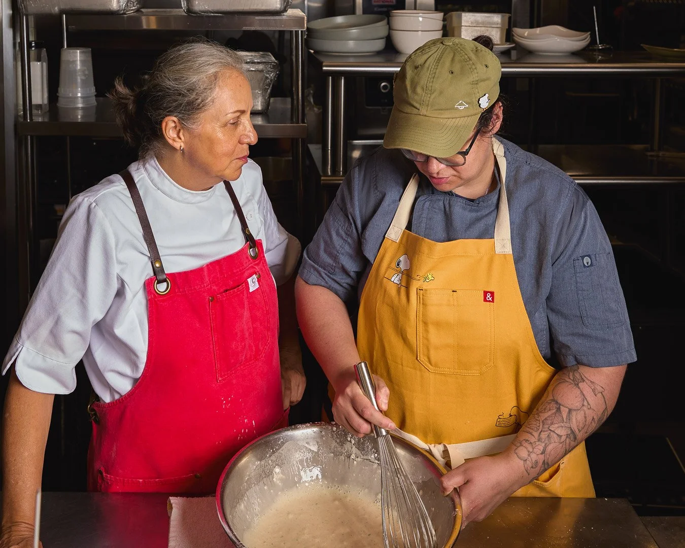 A woman in a red apron is watching a man in a yellow apron and a tan cap who is whisking batter in a bowl in a professional kitchen.