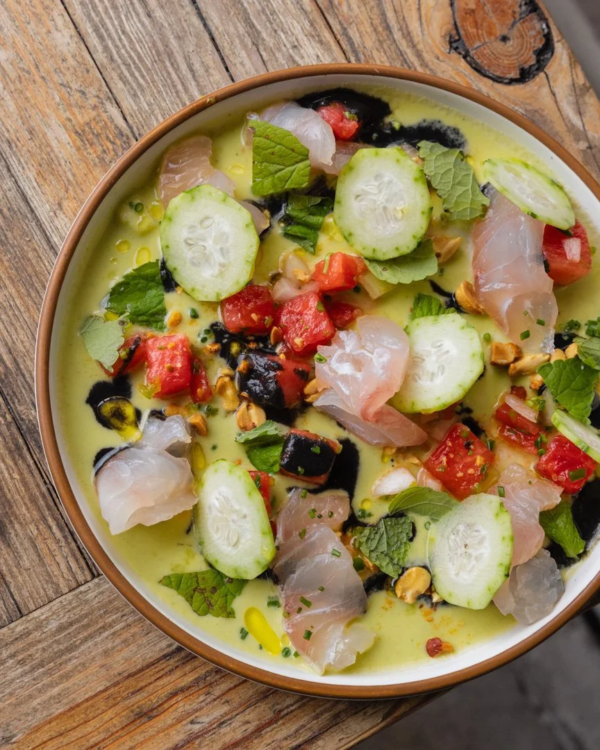 Close-up of a bowl of ceviche with sliced cucumbers, diced tomatoes, cilantro, chopped nuts, black sauce, and oil on a rustic wooden surface.