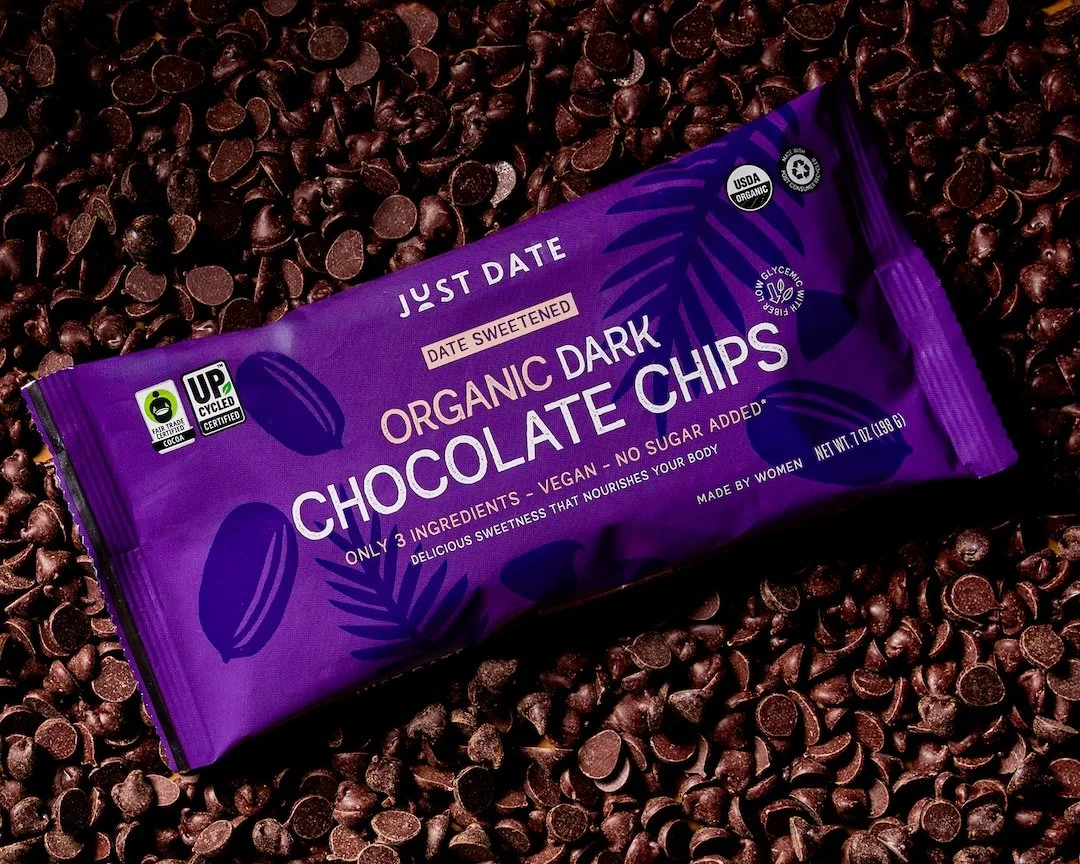 A purple package of Just Date Organic Dark Chocolate Chips resting on a bed of chocolate chips.