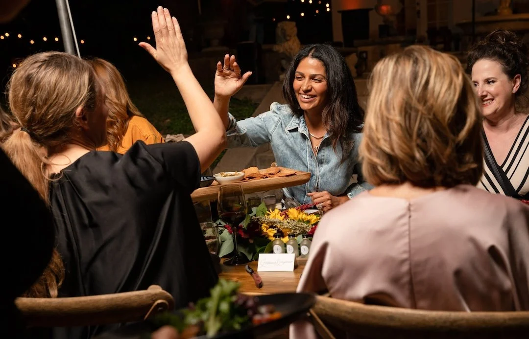Group of women enjoying a dinner party outdoors at night, one woman giving a high five, with food and wine on the table.