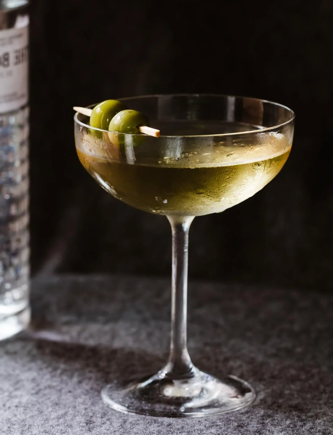 A cocktail glass filled with a light yellow-green drink, garnished with two green olives on a skewer, placed on a dark surface with a black background.