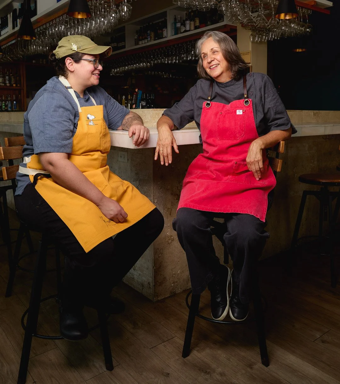 Two women having a conversation in a bar or restaurant, both wearing aprons, one in yellow and the other in red. They are smiling and sitting at a counter, with glasses hanging above them in the background.