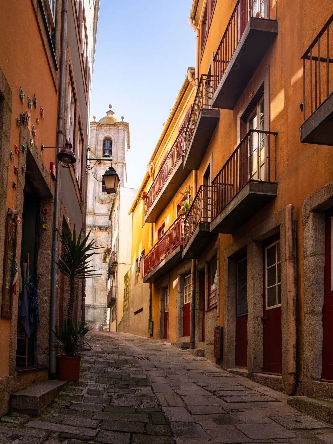 A narrow cobblestone street lined with colorful European-style buildings, potted plants, and a church tower visible in the background under a clear blue sky.