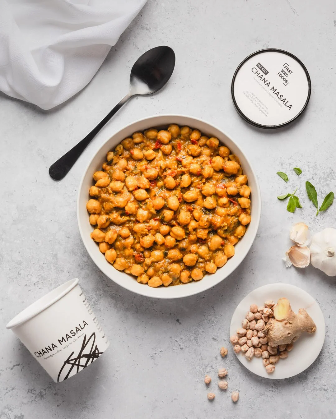A bowl of chickpea curry on a light grey surface, with a black spoon, a container of Chana Masala spice, garlic, ginger, fresh herbs, and a white paper cup labeled 'Chana Masala' around it.