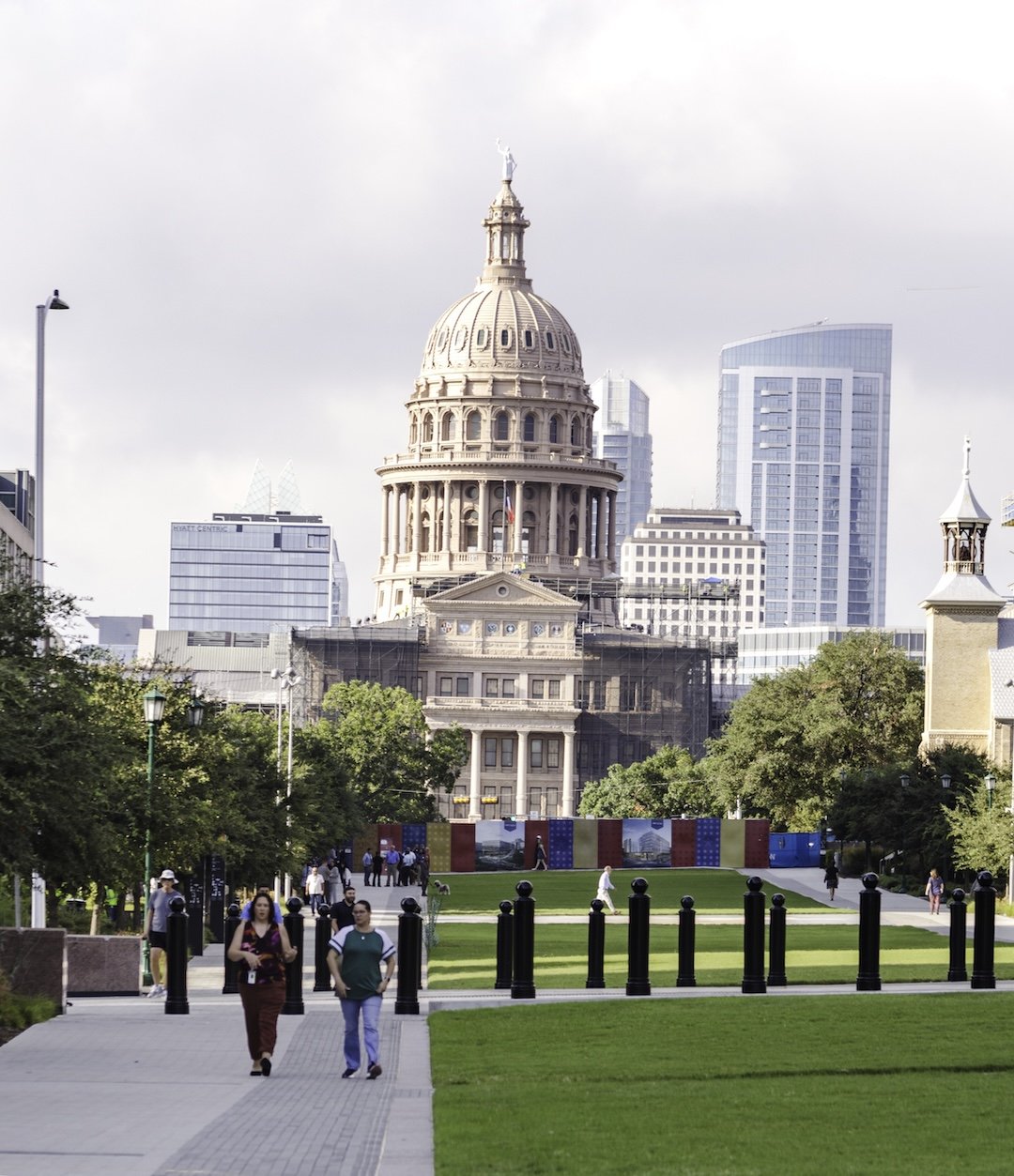 The Texas State Capitol building in Austin with modern skyscrapers in the background, surrounded by a park with people walking and green grass.