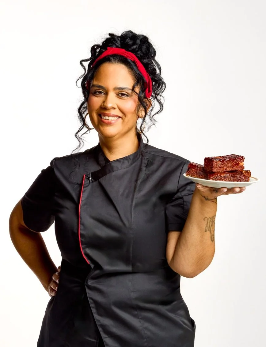 A woman dressed in black cooking attire holding a plate of slab-style ribs with a white background.