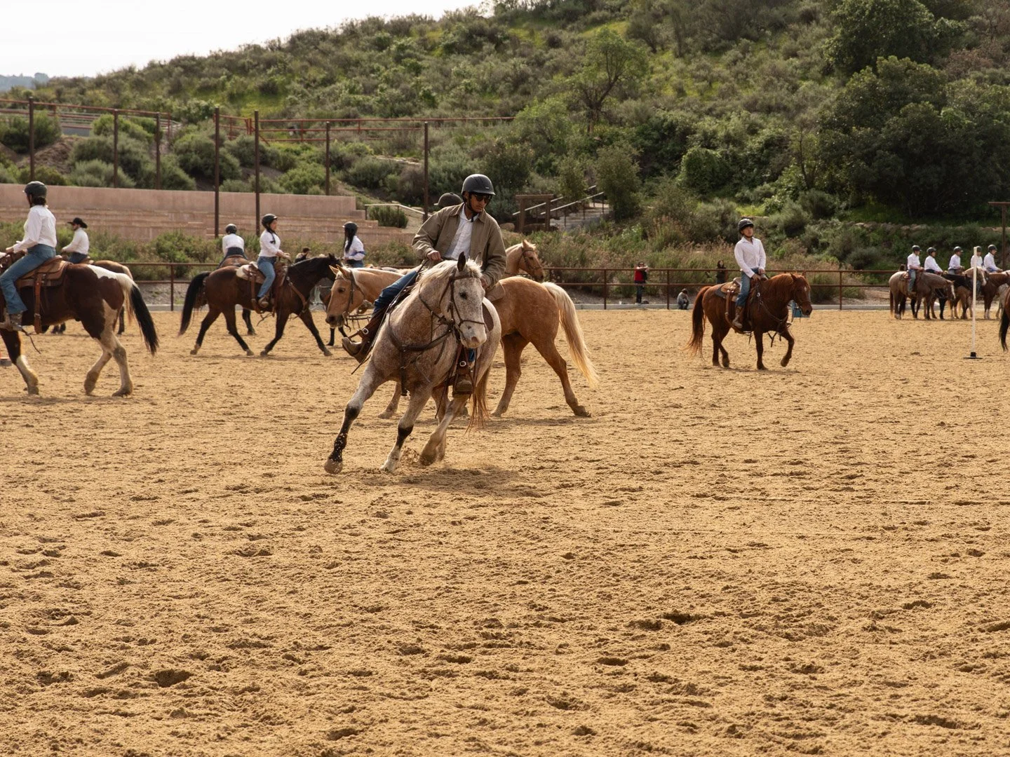 People horse riding on an outdoor arena with sandy ground, surrounded by a fence, with a hill and green trees in the background.
