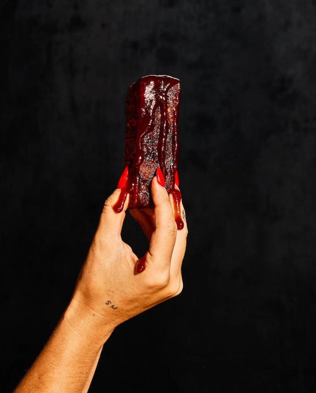 A hand with red-painted nails holding a chocolate-covered ice cream bar with melted red syrup, against a black background.