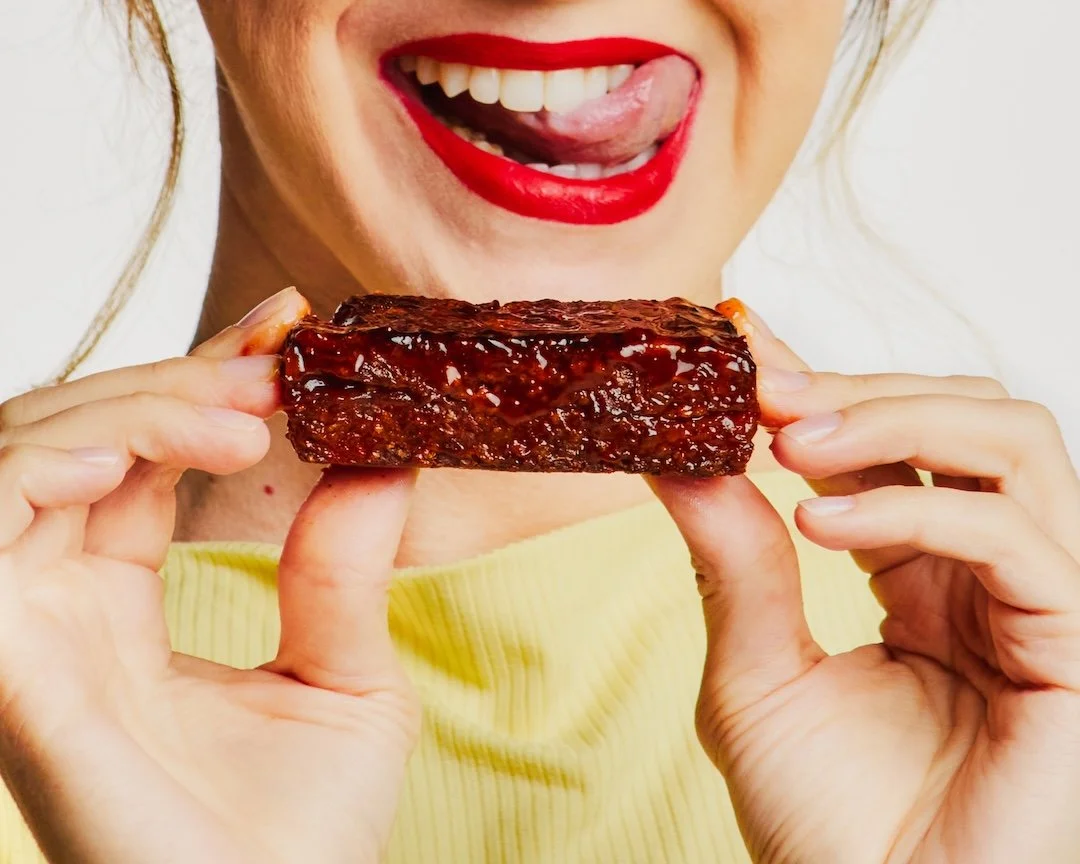 Close-up of a woman smiling with red lipstick, holding a piece of glazed barbecue ribs, licking her lips.