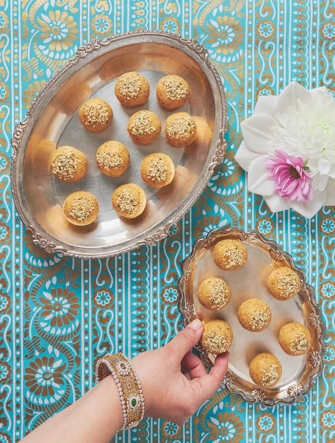 Tray of nine round, gold-colored desserts topped with crushed nuts, placed on an ornate silver tray, with another similar tray held by a hand, on a blue and gold patterned tablecloth, with a white and pink flower nearby.