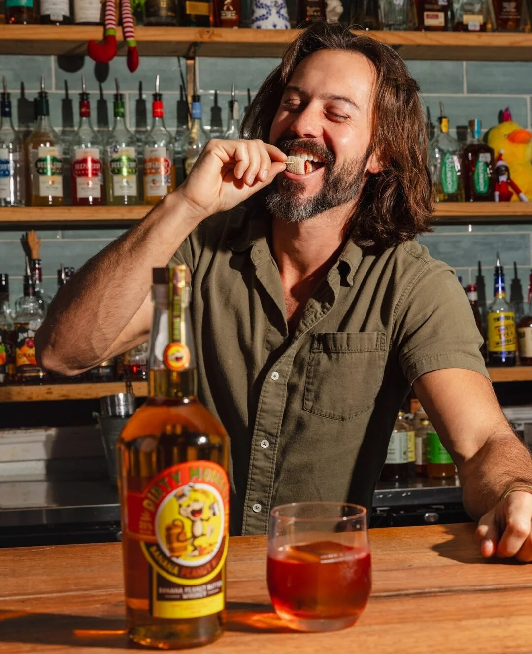 A man with long hair and a beard enjoying a snack at a bar, with a bottle of Hell or High Water beer and a glass of pinkish drink in front of him, shelves of liquor bottles in the background.