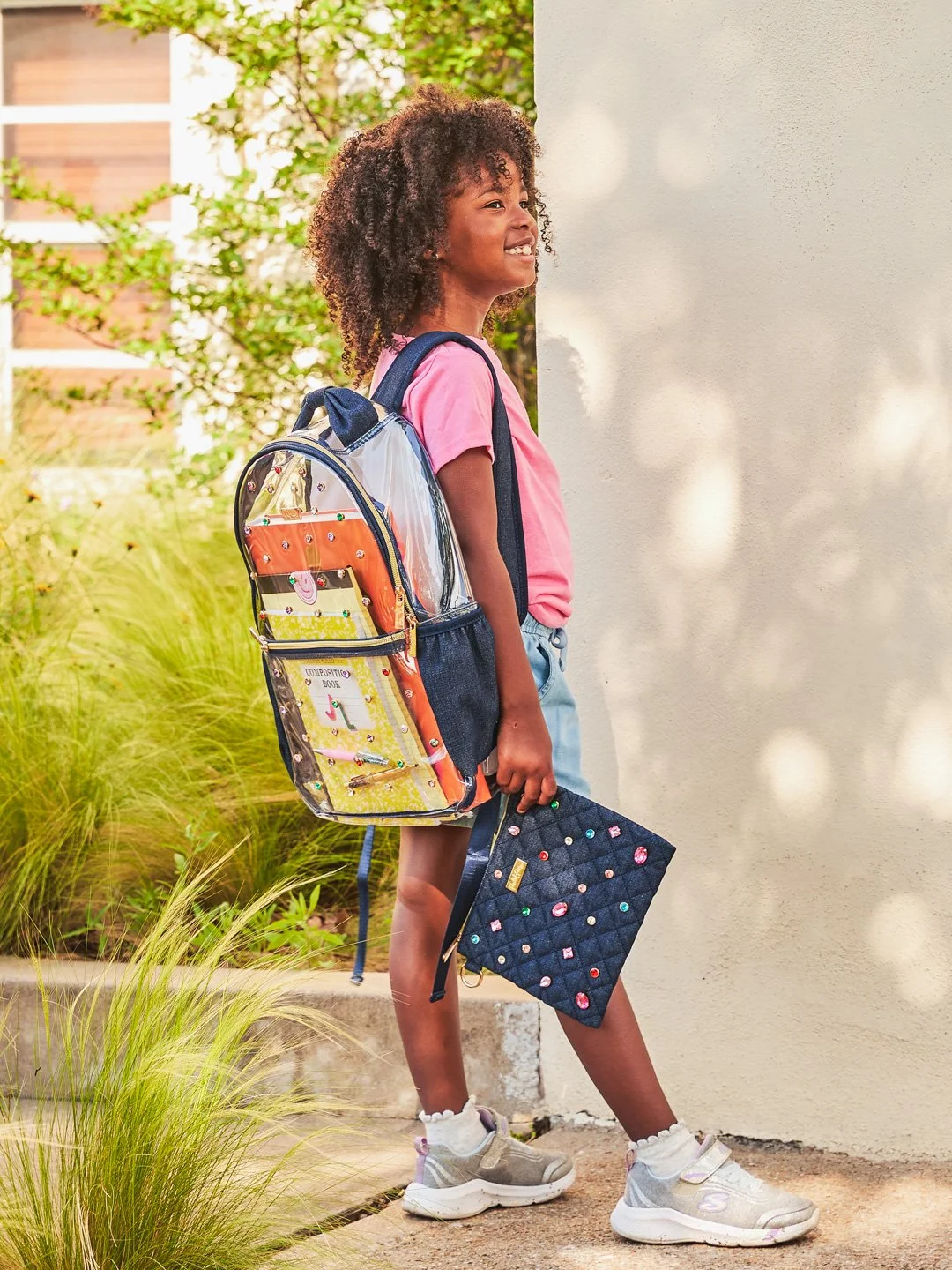 A young girl with curly hair, wearing a pink shirt, shorts, and sneakers, standing outdoors with a backpack and holding a quilted tablet or laptop sleeve, smiling and looking to the side.