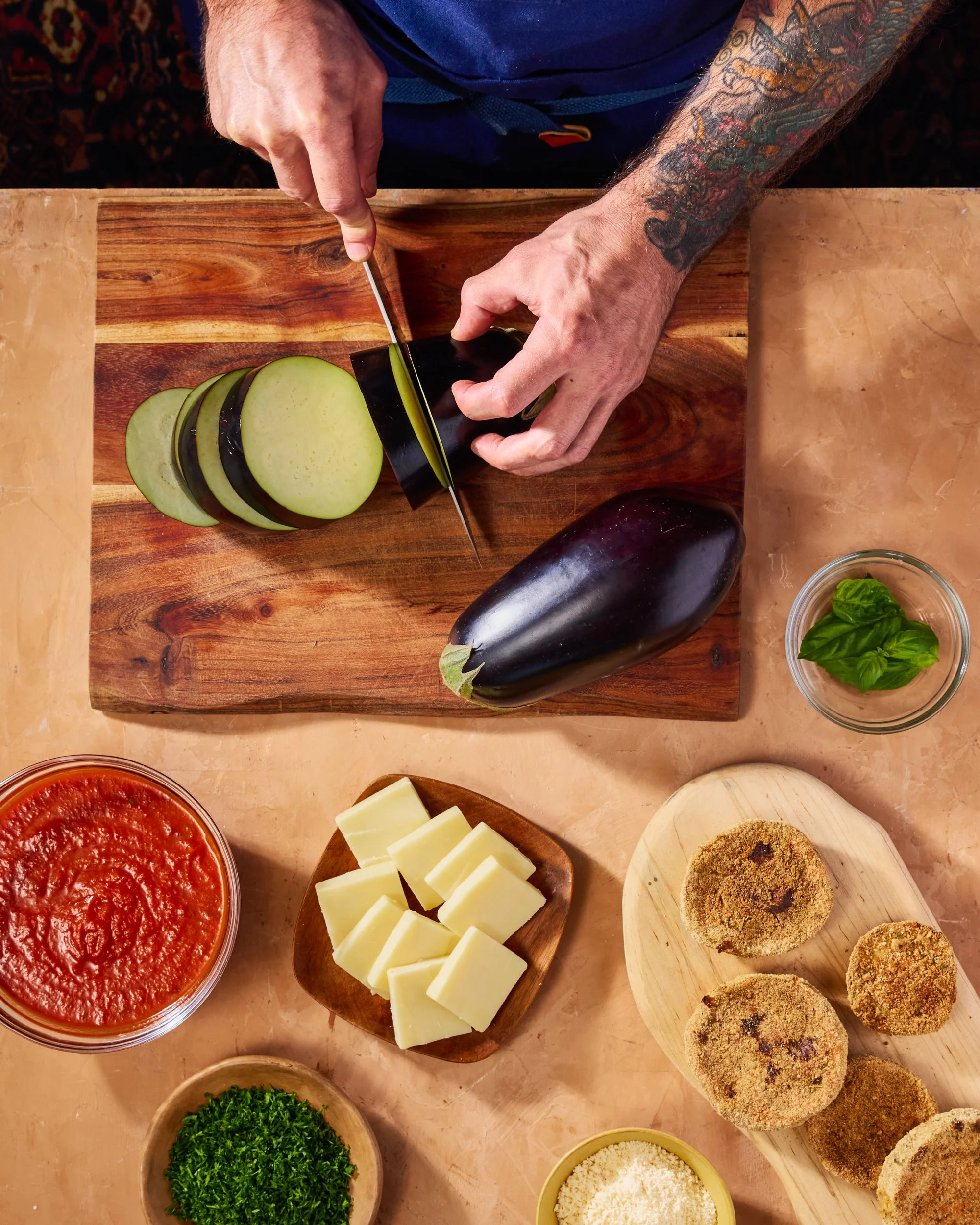 The image shows a person with tattoos on their arms slicing an eggplant on a wooden cutting board. Surrounding the cutting board are bowls of tomato sauce, chopped cheese, fresh green herbs, grated cheese, and breaded items.
