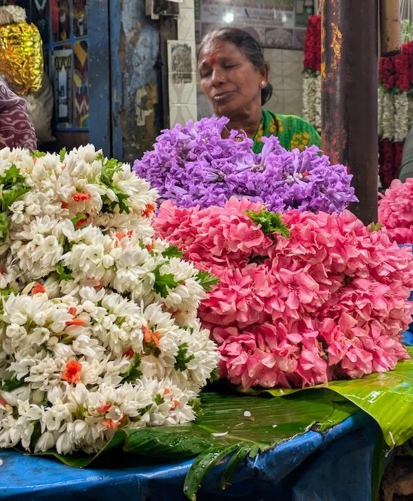 A woman selling large bouquets of white, purple, and pink flowers at an outdoor market.