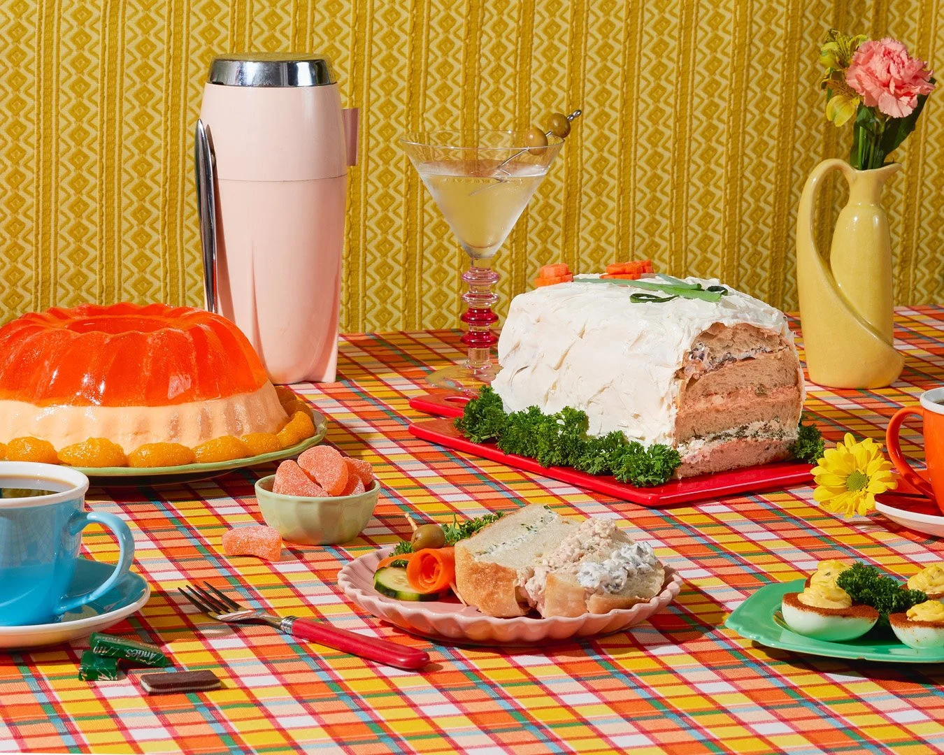 A table set for a meal with a checkered tablecloth, featuring a large decorated meatloaf, a layered cake, a martini glass with a yellow drink, a pink milkshake, and various dishes and cups, with a yellow patterned curtain in the background.