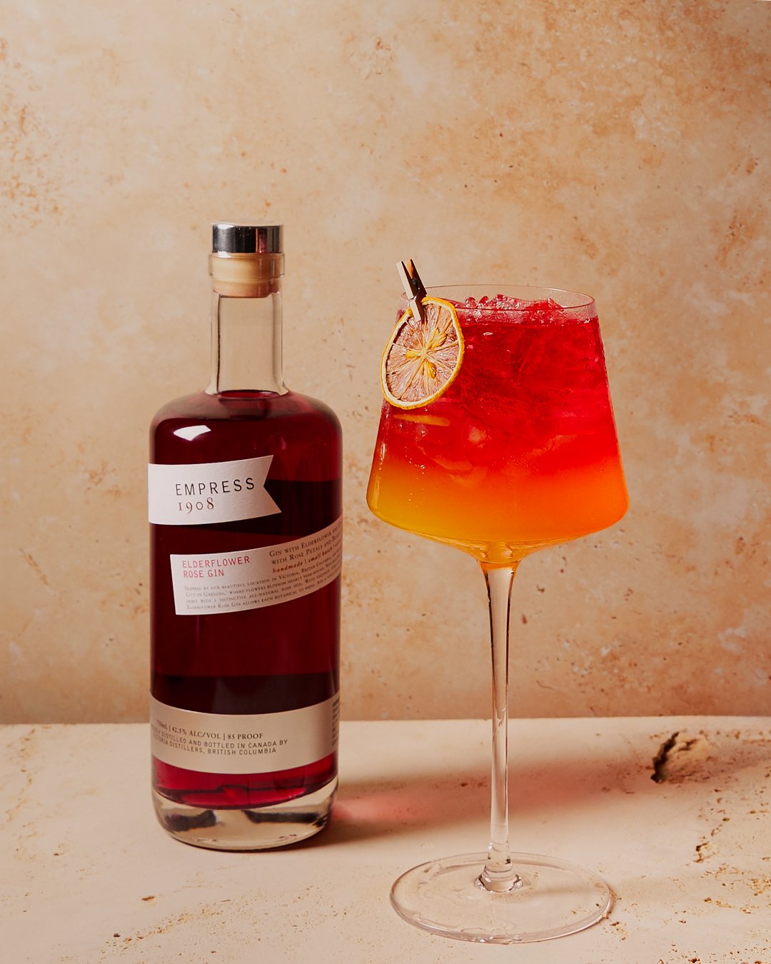Bottle of Empress Elderflower Rose Gin and a colorful cocktail garnished with a dried lemon slice on a stemmed glass with ice and a clothespin, set against a beige textured background.