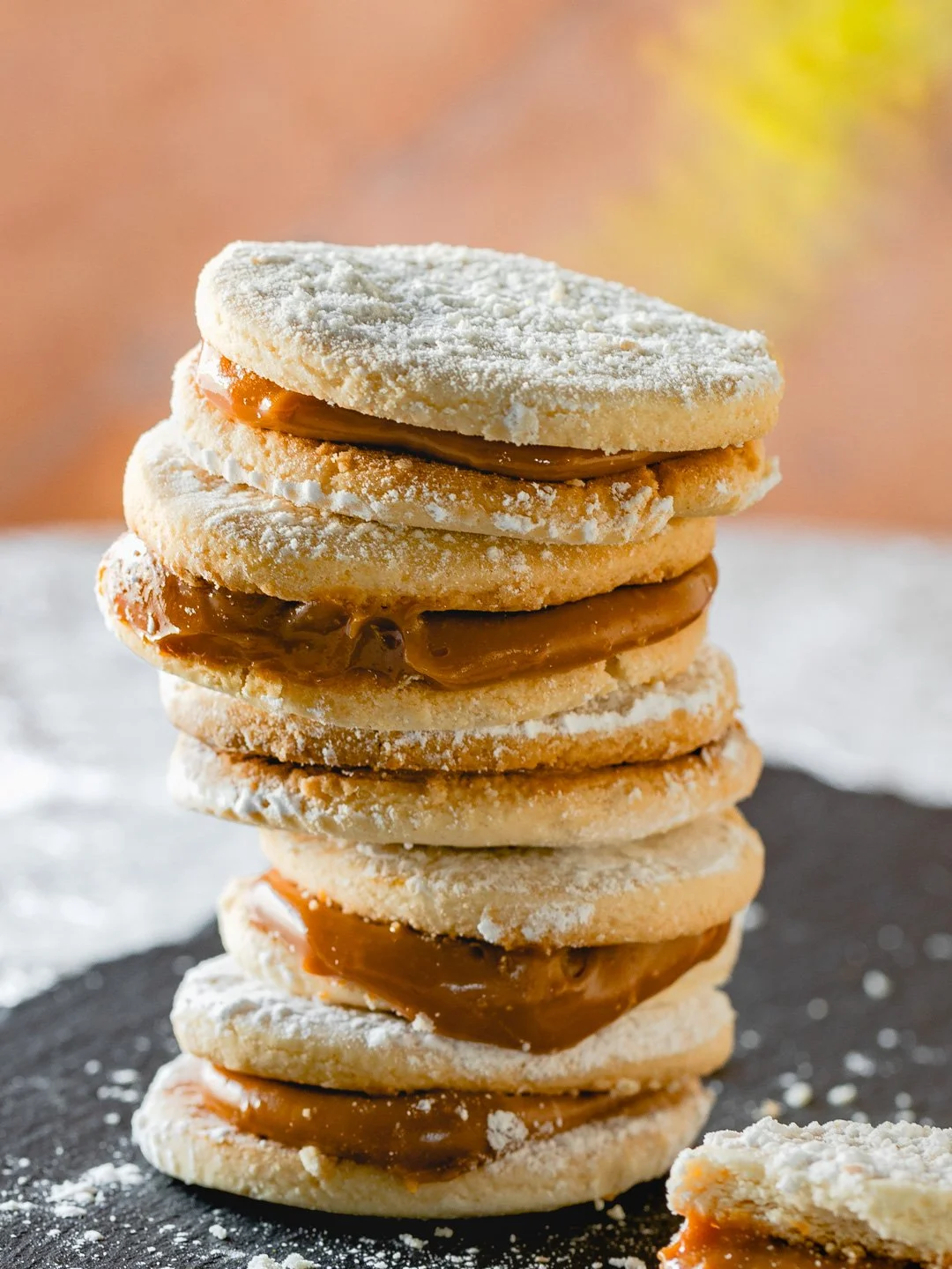 Stack of six thumbprint cookies filled with caramel, dusted with powdered sugar, on a black surface with a blurred autumn background.