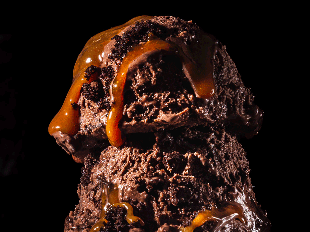 Two scoops of chocolate ice cream topped with caramel sauce and crushed cookie crumbs against a black background.