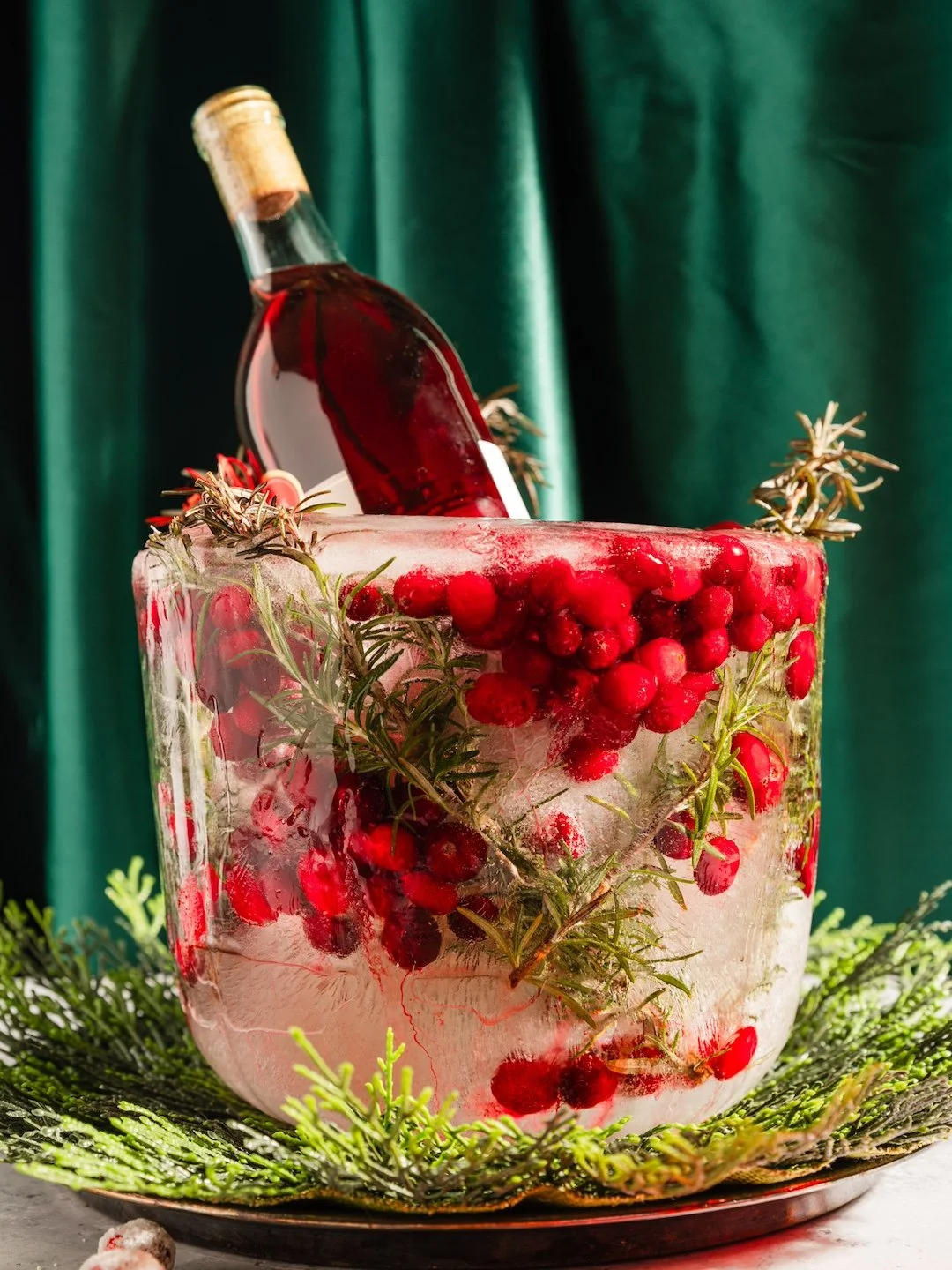 A bottle of red wine in an ice bucket decorated with red berries and fresh rosemary, on a festive green backdrop