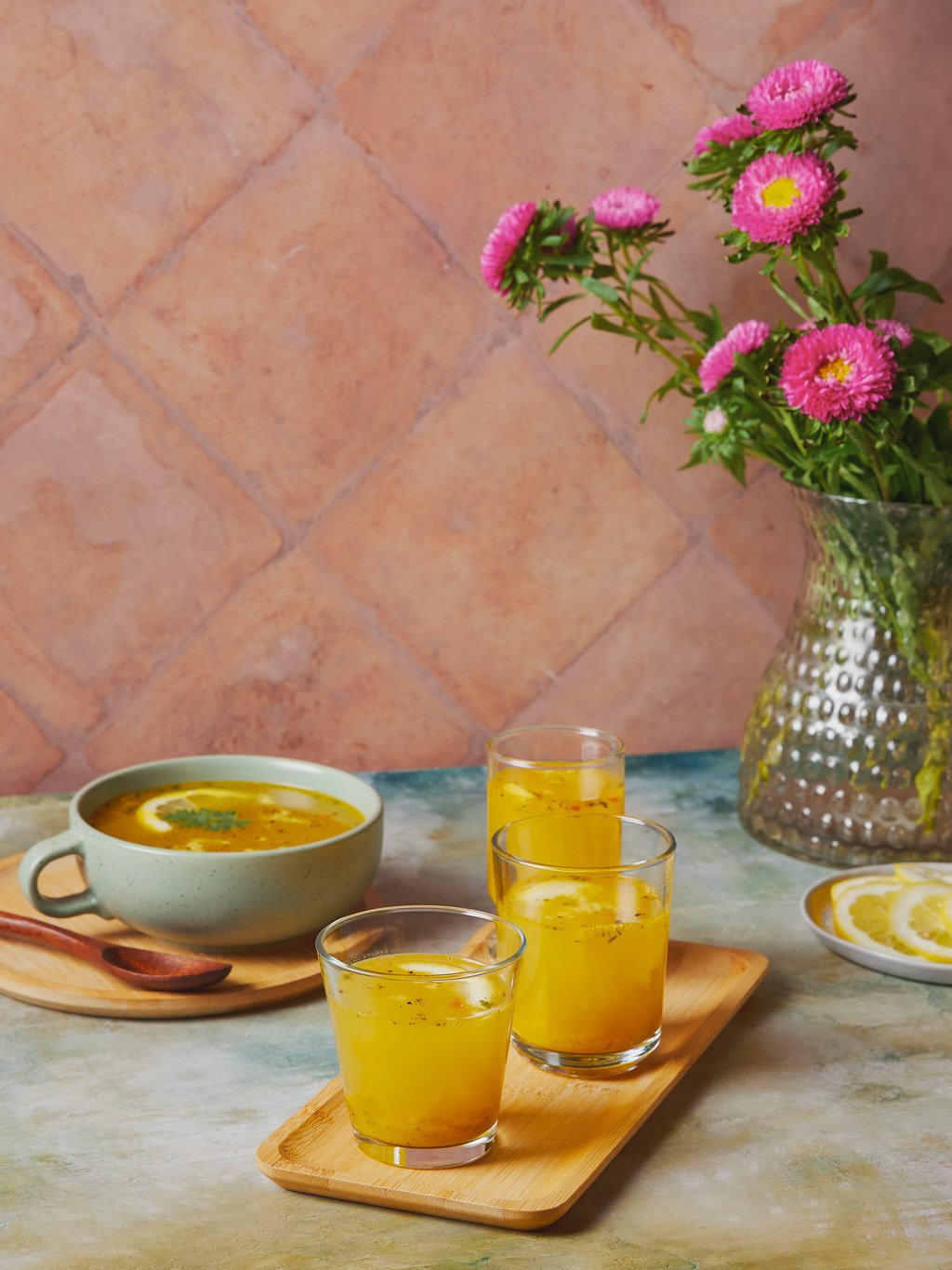 A table with a bowl of soup, three glasses of yellow beverage, a plate with lemon slices, and a vase with pink flowers against a tile wall.