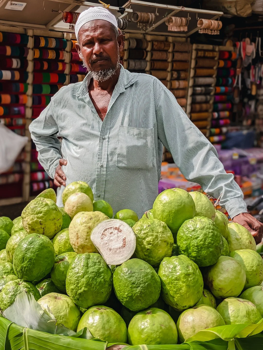 Man standing behind a pile of green and white citrus fruits at a market, with colorful fabric rolls in the background.