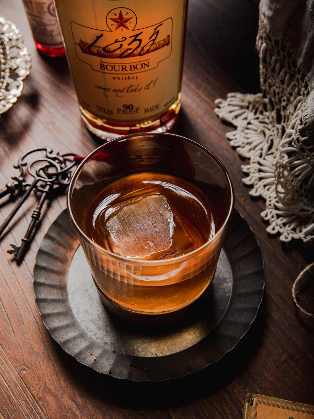 A glass of whiskey with a large ice cube on a metal coaster, with a bottle of bourbon whiskey behind it on a wooden table, decorated with vintage keys, a lace cloth, and a small jewelry box.