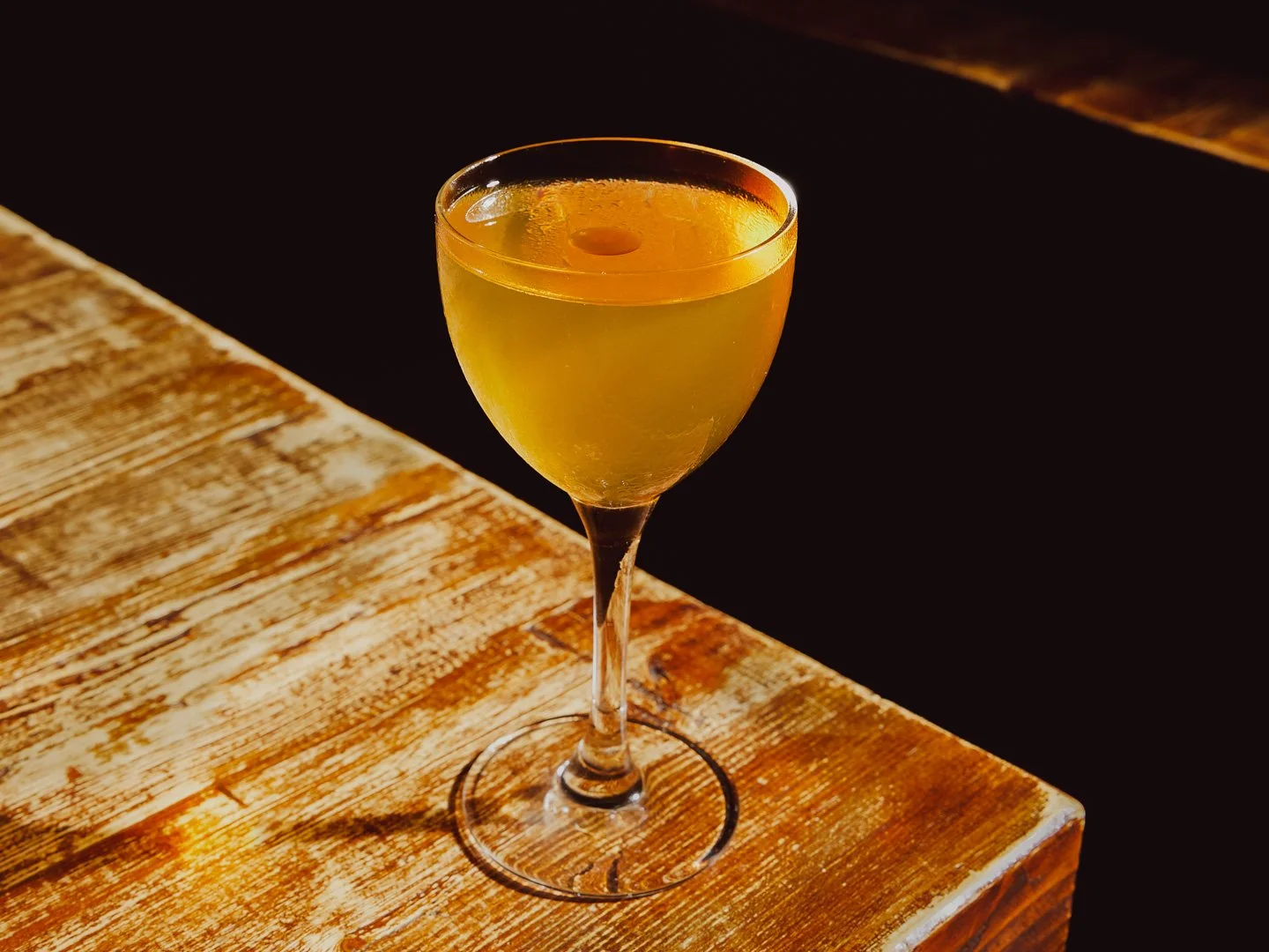 A yellow cocktail in a tall, elegant glass on a wooden table with a dark background.