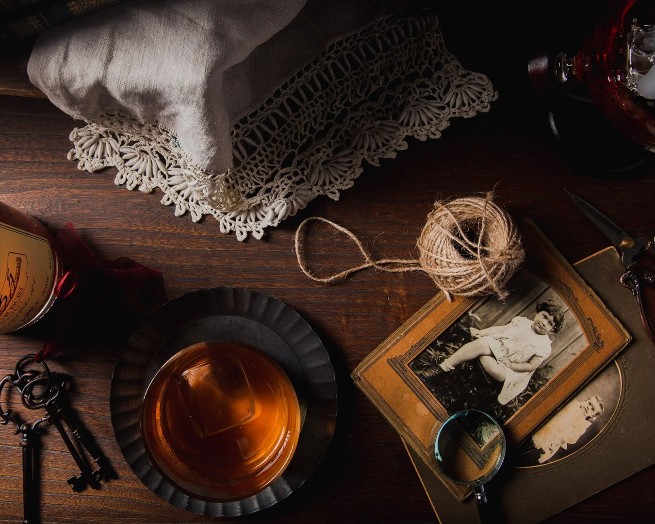 Vintage desk with old photographs, a glass of whiskey, keys, a ball of twine, a lace-trimmed cloth, and a magnifying glass.
