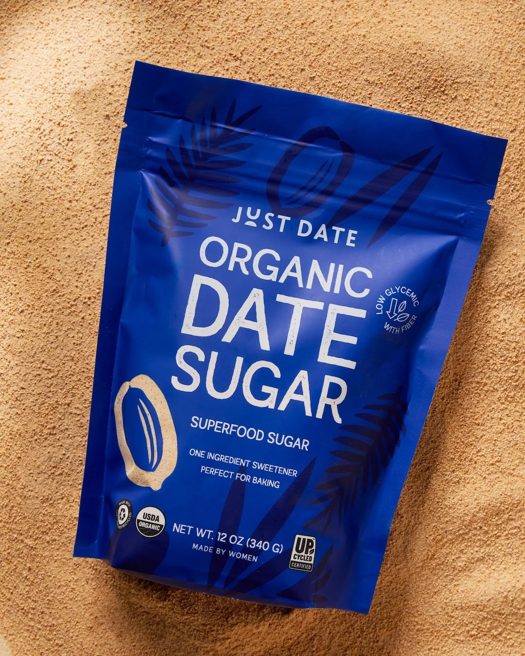 A blue packaging bag of organic date sugar with a palm leaf design, stating 'Just Date Organic Date Sugar,' net weight 12 oz, made by women, and certified organic and USDA organic.
