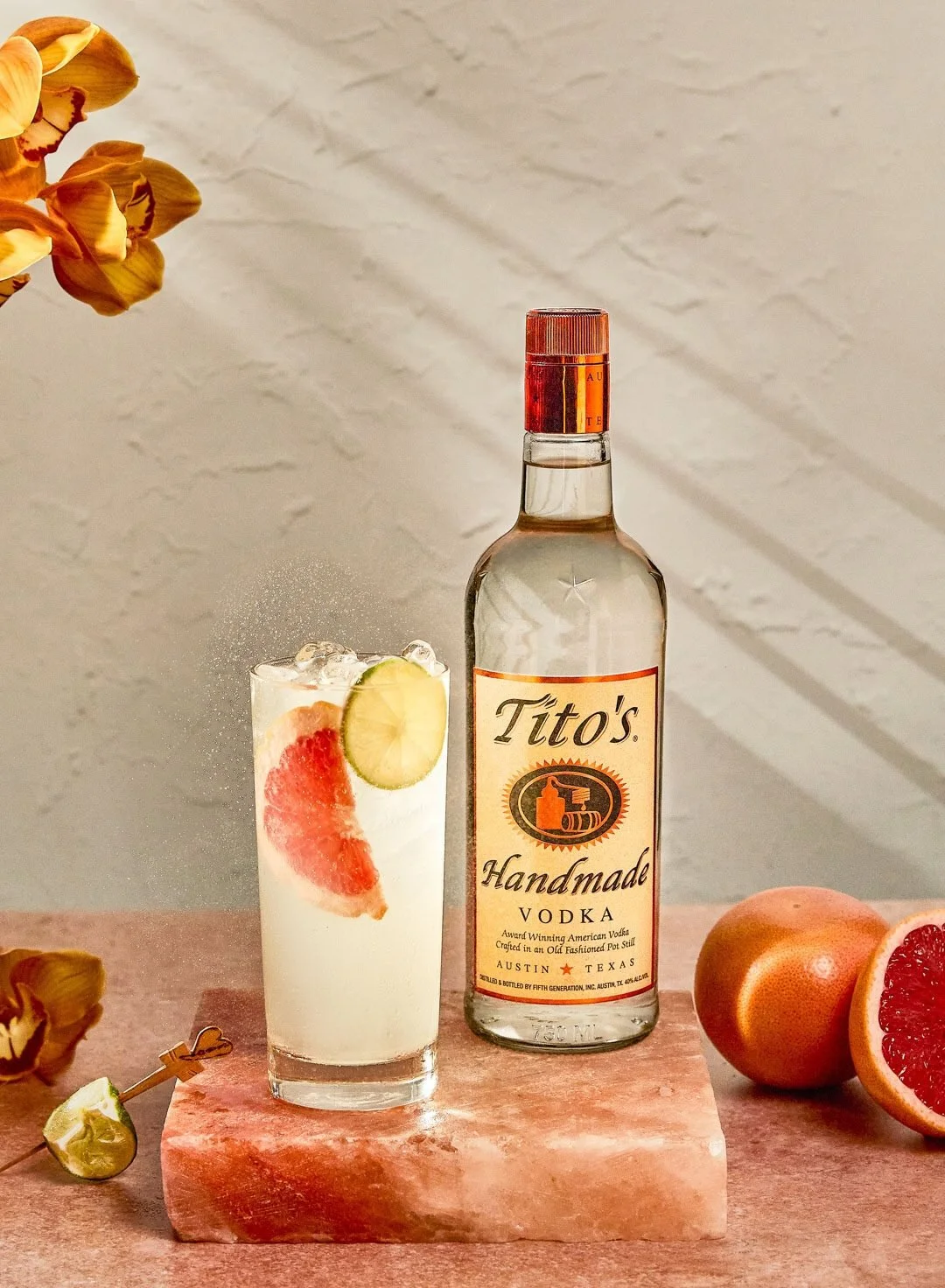 A bottle of Tito's Handmade Vodka beside a glass with a grapefruit slice, a lime wedge, and ice, on a pink marble surface surrounded by oranges and flower petals.