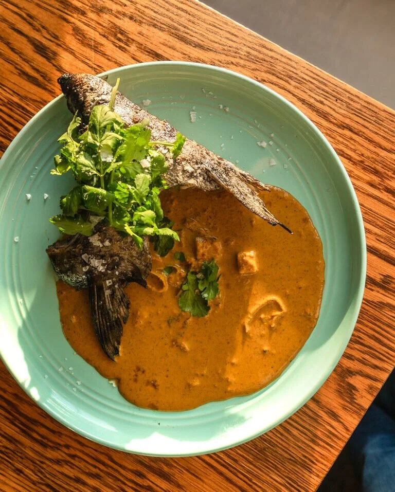 A green bowl containing a fish head in a spicy sauce, garnished with fresh cilantro and some greens, placed on a wooden table.