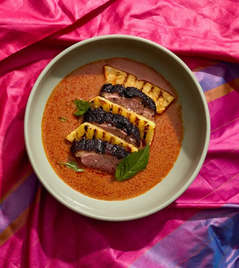 Sliced grilled steak with caramelized pineapple and basil leaves in a bowl of tomato-based sauce.