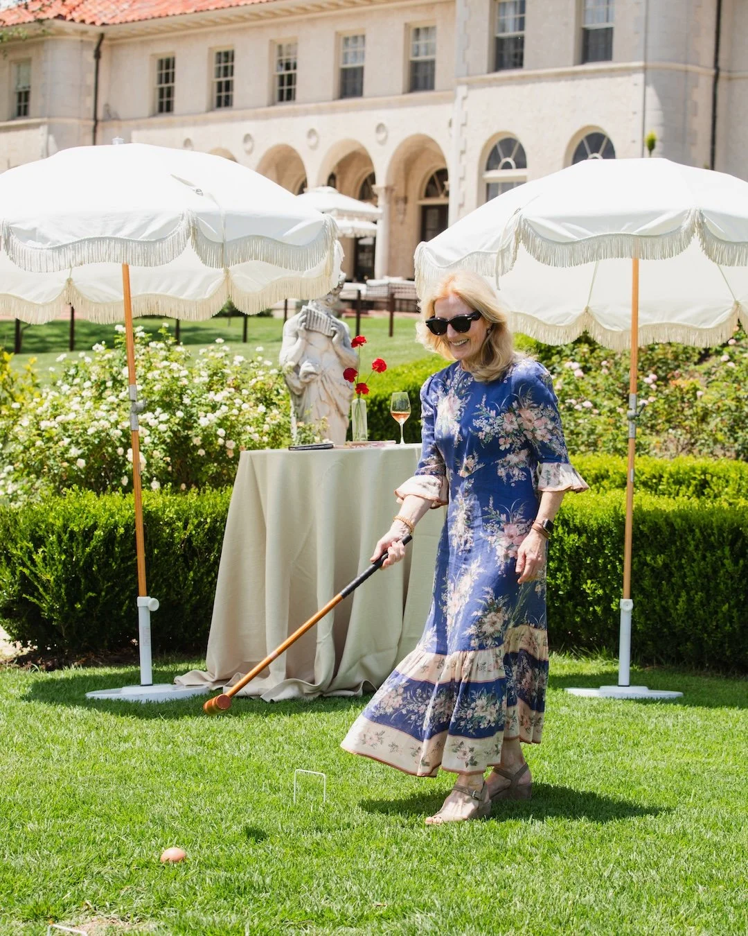 Woman in floral dress playing croquet on a lawn with umbrellas, a table with wine and flowers, and a building in the background.