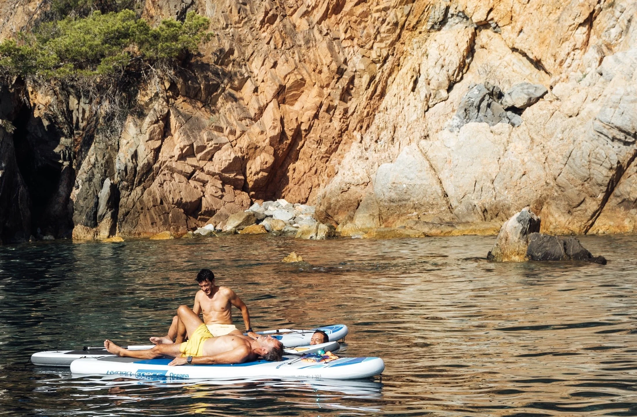 A man lying on a paddleboard in water, with another man sitting on his lap, both enjoying a sunny day near a rocky cliff.