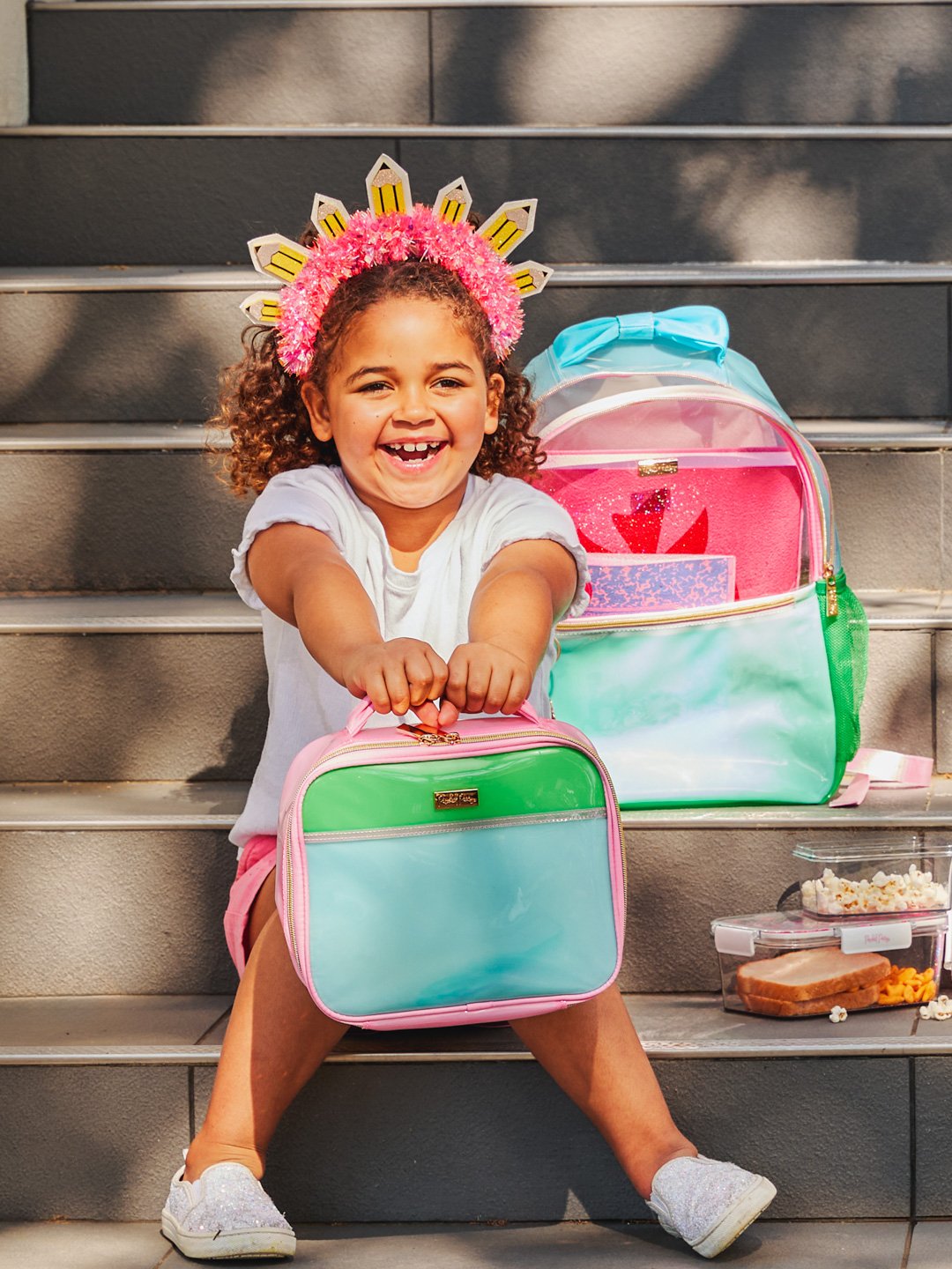 A smiling young girl with curly hair wearing a pink and white crown, sitting on stairs, holding a pastel-colored lunch bag, with a larger matching backpack and a snack container beside her.