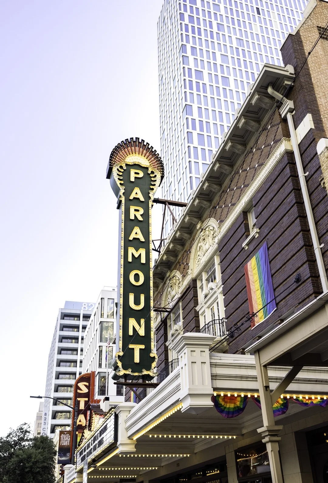 The Paramount Theatre marquee sign with the name vertically displayed next to a tall skyscraper in the background. A rainbow pride flag is hanging on the building, and there is decorative rainbow bunting under the awning.