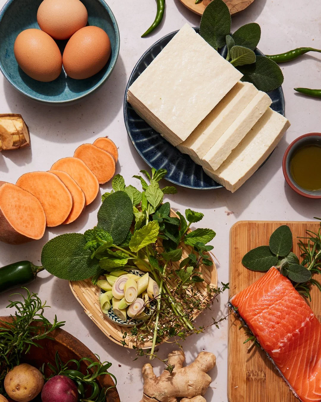 Eggs in a blue bowl, sliced sweet potatoes, fresh ginger, various fresh herbs like sage and mint, sliced green chili, raw salmon fillet on a wooden cutting board, small bowl of olive oil, green chili, and a variety of vegetables including potatoes an