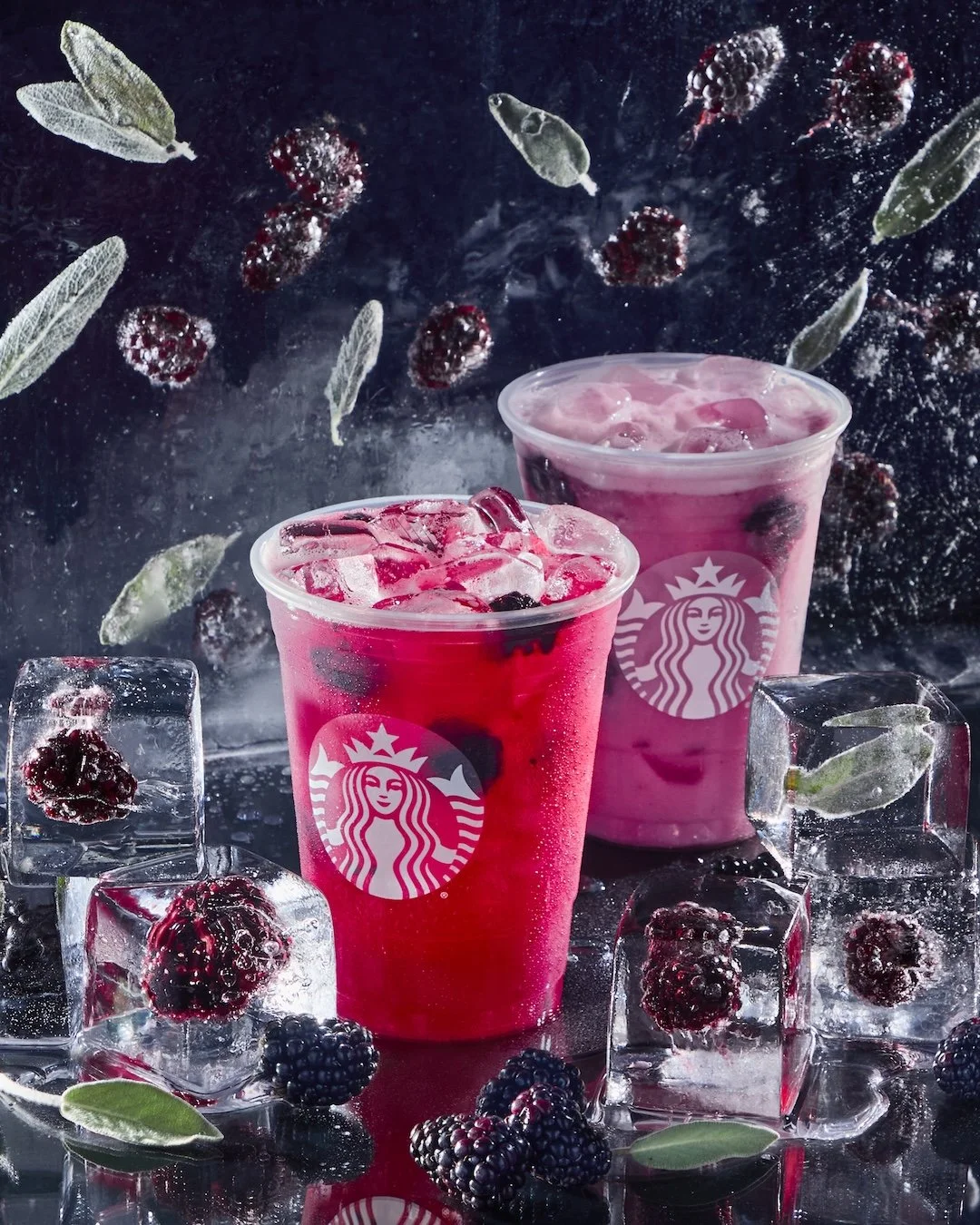 Two Starbucks beverages, one pink and one purple, surrounded by ice cubes with blackberries, red berries, and green leaves inside and around, on a reflective surface with a dark backdrop and floating berries and leaves.