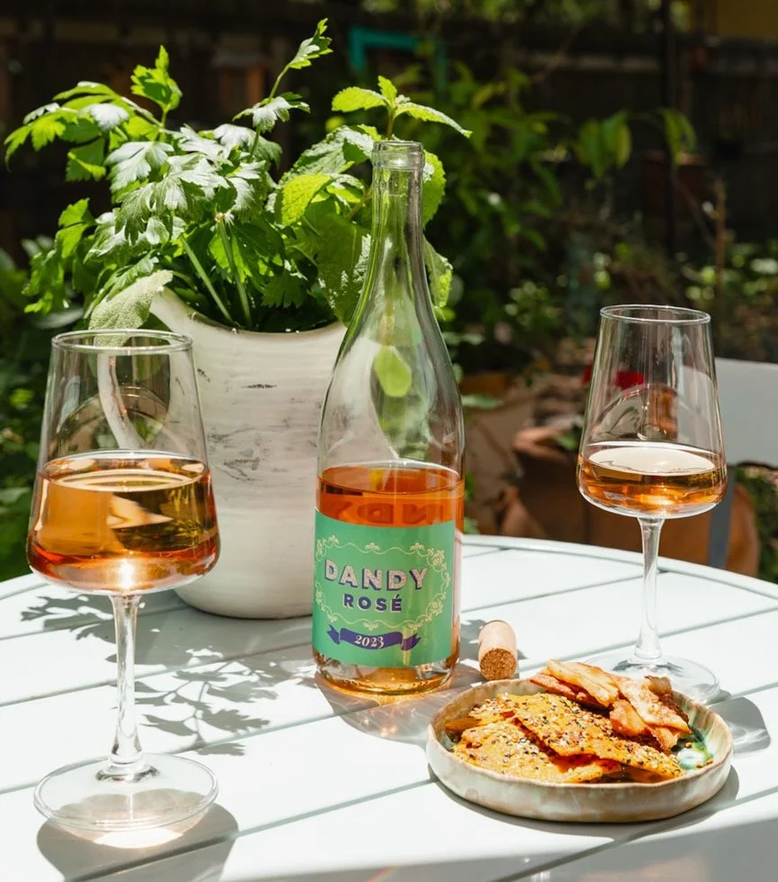 A bottle of Dandy Rosé 2023 wine, two glasses of rosé wine, a plate of crackers or bread, a potted green plant, and a cork on a white outdoor table in a garden setting.