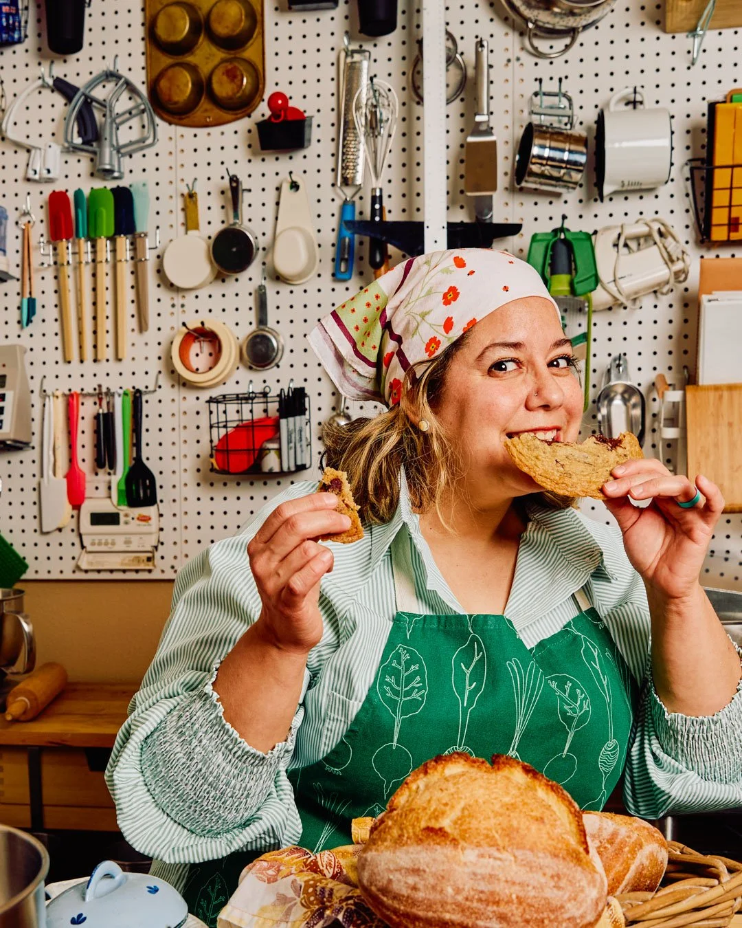 A woman wearing an apron and a floral headscarf, smiling and eating a slice of bread in a kitchen with utensils hanging on a pegboard behind her.