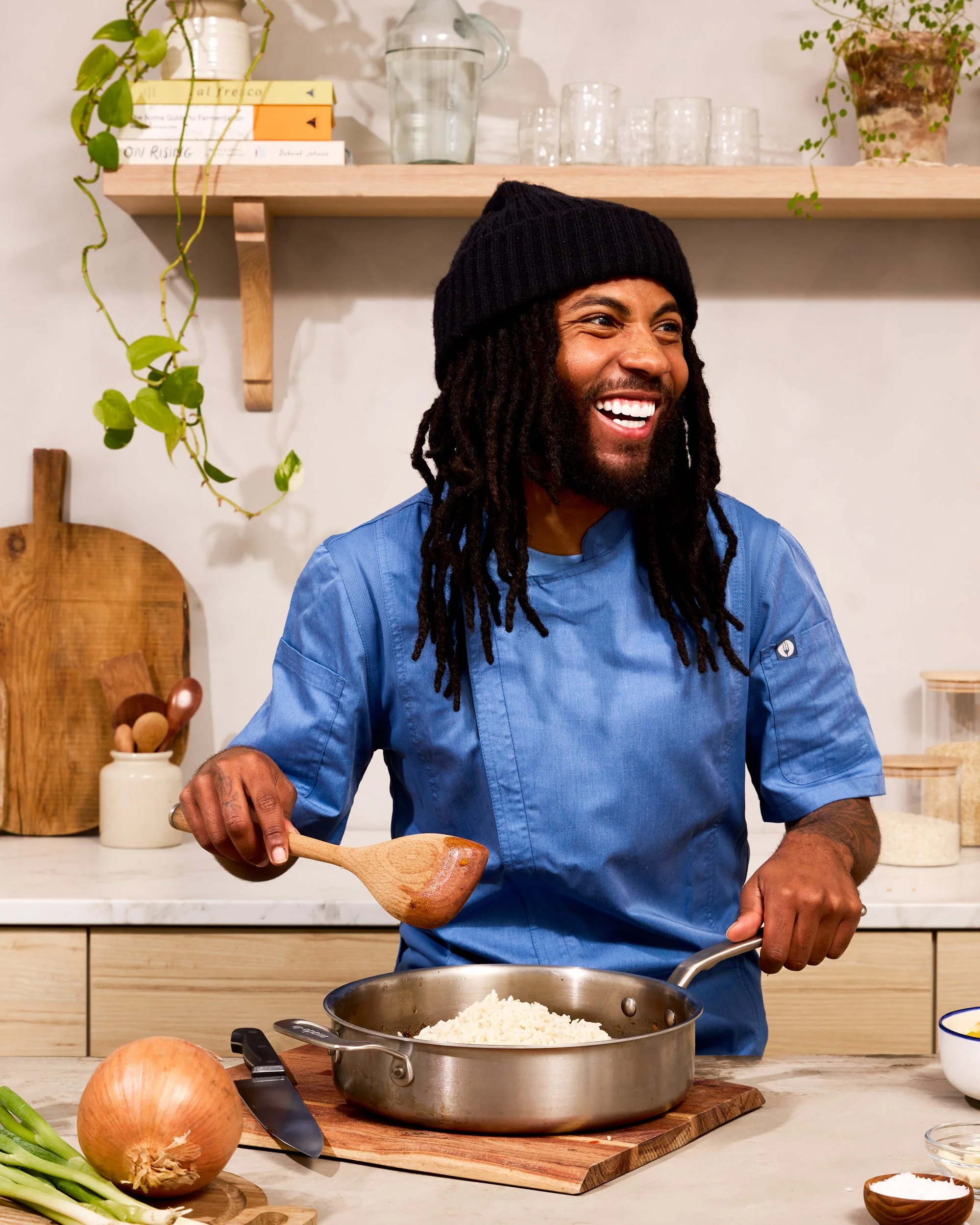 Man with dreadlocks, black beanie, and blue chef's coat cooking rice in a sauté pan in a modern kitchen with wooden shelves, potted plants, and cooking ingredients.
